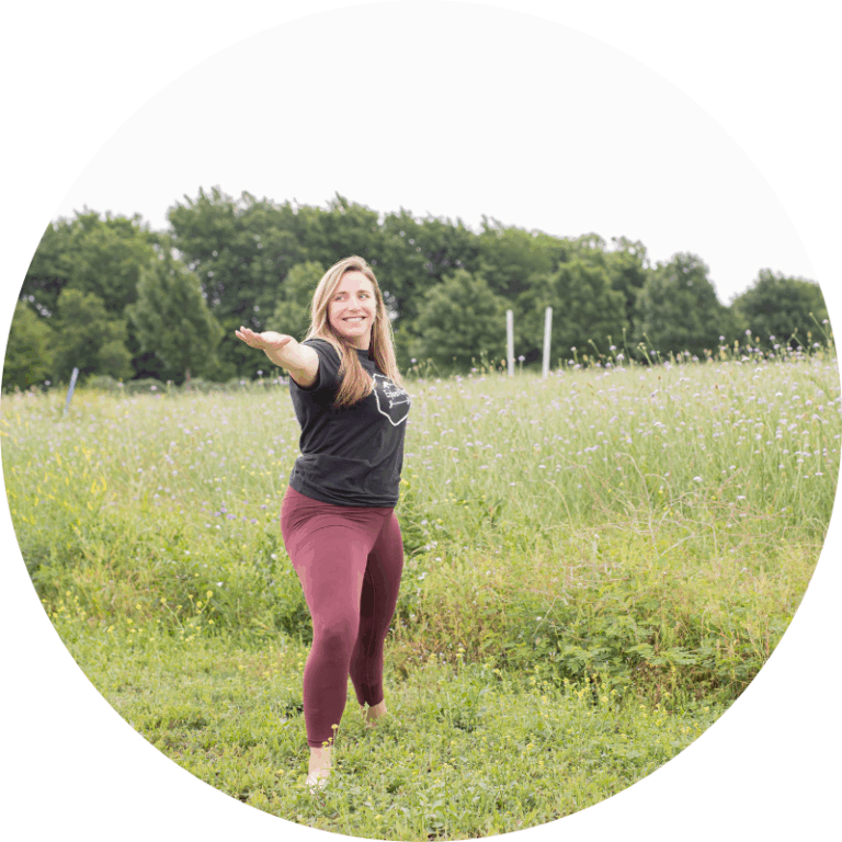 Haley Cottey in Warrrior 2 shape in a field of grass with trees in the distance